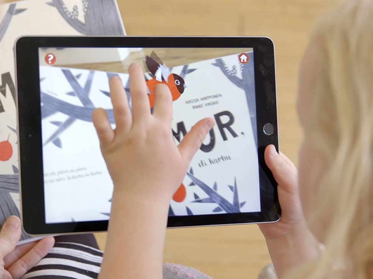 A child holds an iPad over the picture book Mur; the bear appears on the screen